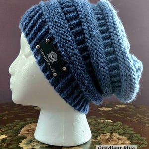 May include: A gradient blue knit beanie hat. The hat is displayed on a white mannequin head. The hat has a dark blue band with a black label that reads "SLIP. STITCH. SNIP" with a skull and crossbones logo. The hat is labeled "Gradient Blue".