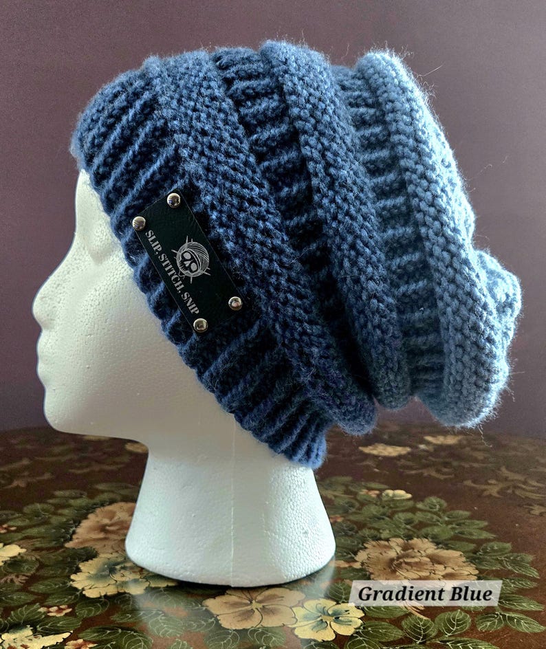 May include: A gradient blue knit beanie hat. The hat is displayed on a white mannequin head. The hat has a dark blue band with a black label that reads "SLIP. STITCH. SNIP" with a skull and crossbones logo. The hat is labeled "Gradient Blue".