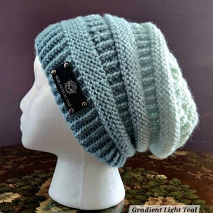 May include: A light teal knitted beanie hat with a gradient effect. The hat features a textured stitch pattern and a black label with white text that reads "SLIP.STITCH.SNIP". The hat is displayed on a white mannequin head.