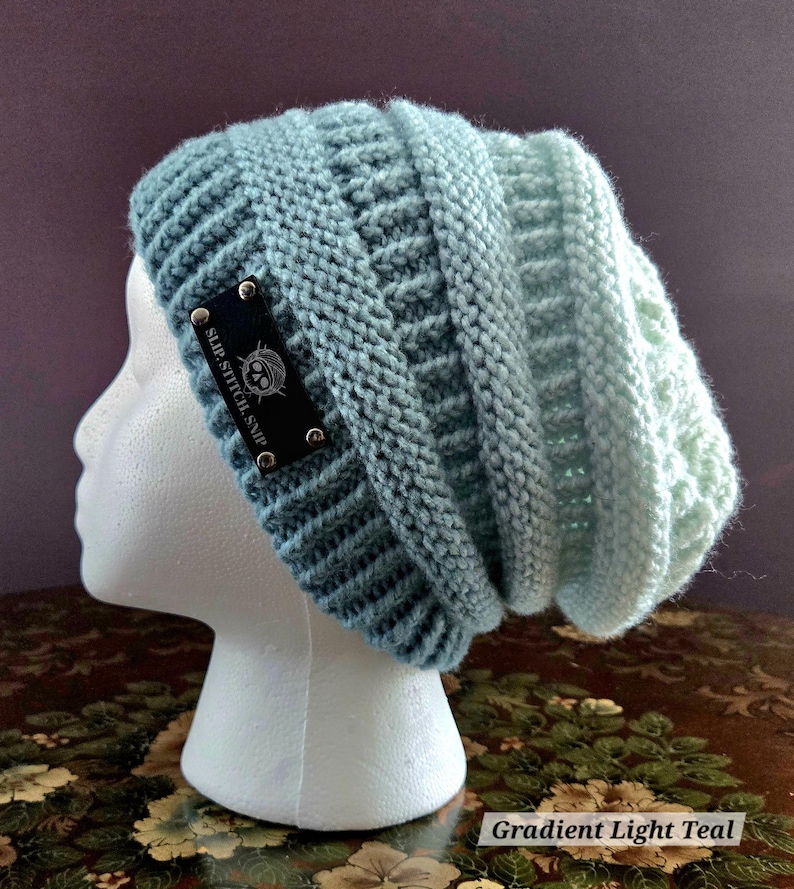 May include: A light teal knitted beanie hat with a gradient effect. The hat features a textured stitch pattern and a black label with white text that reads "SLIP.STITCH.SNIP". The hat is displayed on a white mannequin head.