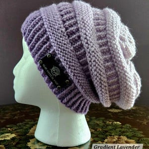May include: A gradient lavender knit beanie with a textured design. The hat features a dark label with the text "SLIP.STITCH.SNIP" and a skull logo. The beanie is displayed on a white mannequin head.