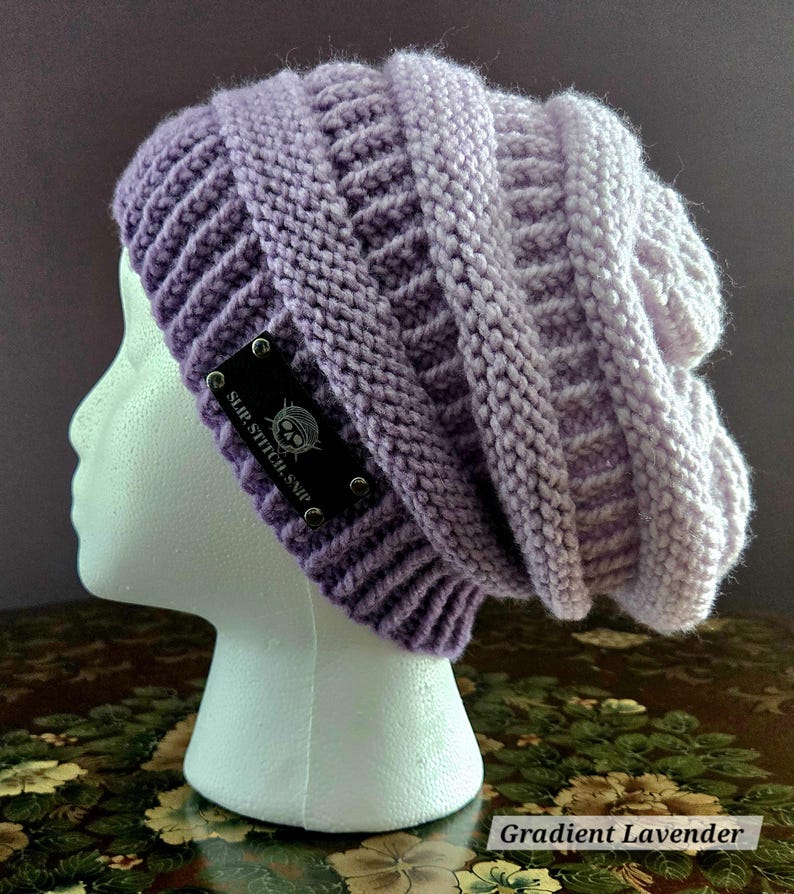May include: A gradient lavender knit beanie with a textured design. The hat features a dark label with the text "SLIP.STITCH.SNIP" and a skull logo. The beanie is displayed on a white mannequin head.