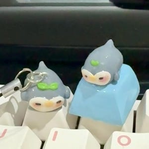 May include: Two gray penguin keycaps with green bow accents and a keychain. One penguin is on a blue keycap, the other is on a white keycap. The keycaps are on a white keyboard.