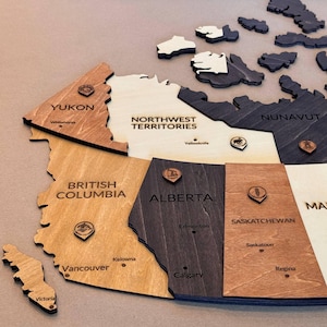 Peut inclure: Une carte murale en bois du Canada présentant des provinces et territoires découpés au laser dans des tons de bois clair, moyen et foncé. Chaque région est étiquetée avec son nom et ses grandes villes, avec de petites icônes en bois.