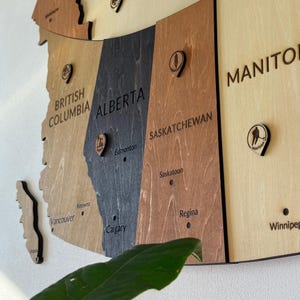 May include: A wooden map of Canadian provinces, with each province in a different wood stain colour. The provinces of British Columbia, Alberta, Saskatchewan, and Manitoba are visible, with city names and decorative pins.