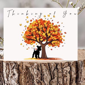 Puede incluir: Una tarjeta de felicitación con el texto "Thinking of You" presenta una ilustración de un árbol con hojas naranjas y amarillas, una silueta de perro negro y hojas cayendo. La tarjeta se muestra sobre una rodaja de madera con un fondo de mármol.