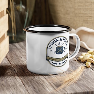 May include: White enamel mug with a black rim and handle. The mug features a blue and gold logo that reads "Union & Brew" with the text "97th Pennsylvania" and the year "1861". The mug is sitting on a wooden surface.