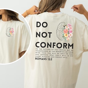 Könnte beinhalten: Cremefarbenes T-Shirt mit dem schwarzen Aufdruck "DO NOT CONFORM". Auf der Rückseite befindet sich eine Gehirngrafik, die mit bunten Blumen gefüllt ist. Der Text "ROMANS 12:2" steht unter einem längeren Zitat.