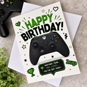 May include: A white birthday greeting card featuring a black video game controller graphic, the text HAPPY BIRTHDAY!, and a green pixelated speech bubble with the text ANOTHER YEAR, NEW LEVEL, YOU'VE GOT THIS! and the text 1UP.