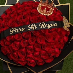 Könnte beinhalten: Ein großer Strauß roter Rosen, verpackt in schwarzem Papier mit goldenen Akzenten. Ein schwarzes Band mit dem goldenen Schriftzug "Para Mi Reyna" ist über den Blumen drapiert. Eine goldene Krone und ein Schmetterling setzen dekorative Akzente.