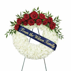 Può includere: Una corona floreale bianca con un nastro blu scuro con la scritta dorata "From the Wilson Family". La corona è adornata con rose rosse e fogliame verde, posta su un supporto in metallo nero.