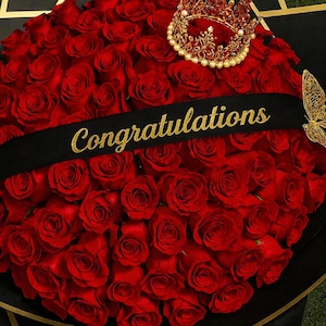 Può includere: Un grande bouquet di rose rosse, avvolto in carta nera con dettagli dorati. Un nastro nero con la scritta "Congratulazioni" in oro attraversa le rose. Una corona e una farfalla dorate completano il tema celebrativo.
