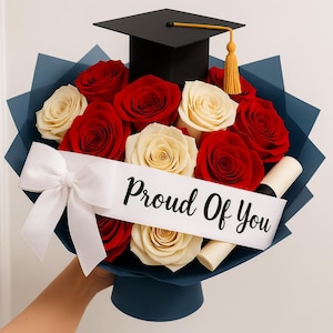 Puede incluir: Un ramo de graduación con rosas rojas y crema, un birrete negro y un pergamino. El ramo está envuelto en papel azul y atado con una cinta blanca que dice "Proud Of You".
