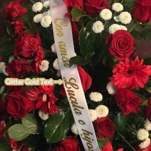 Peut inclure: Un arrangement floral vibrant avec des roses rouges, des chrysanthèmes blancs et des gerberas rouges. Un ruban blanc avec du texte doré à paillettes indiquant "Con amor Lucila e hijos" est drapé sur les fleurs.