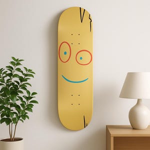 Puede incluir: Una patineta amarilla con un diseño de cara de dibujos animados. La cara presenta ojos delineados en rojo con pupilas azules, una sonrisa azul y detalles de cabello negro. La patineta está montada en una pared blanca.