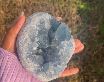 May include: A light blue celestite geode with crystal formations. The geode is held in a person's hand, with a soft purple sleeve visible in the frame. The background is blurred, showing green grass and sunlight.