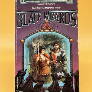 Op de afbeelding: Een paperback boek getiteld "Black Wizards" uit de Forgotten Realms-serie. De cover toont fantasy-kunst met twee figuren, een vrouw en een man, en verschillende figuren met capuchons. Het boek is Boek Twee van The Moonshae Trilogy.