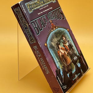 Op de afbeelding: Een paperback boek getiteld "Black Wizards" uit de fantasy-avonturenserie "Forgotten Realms". De cover toont een fantasie-illustratie met figuren in een bosrijke omgeving. Het boek staat op een transparante standaard tegen een gele achtergrond.