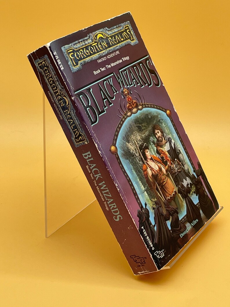 Op de afbeelding: Een paperback boek getiteld "Black Wizards" uit de fantasy-avonturenserie "Forgotten Realms". De cover toont een fantasie-illustratie met figuren in een bosrijke omgeving. Het boek staat op een transparante standaard tegen een gele achtergrond.