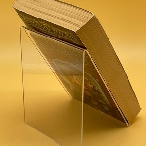 Op de afbeelding: Een heldere acryl boekenstandaard met een hardcover boek. Het boek staat schuin, waardoor de kaft en pagina's zichtbaar zijn. De standaard is transparant, waardoor de kaft van het boek zichtbaar is. De achtergrond is effen geel.
