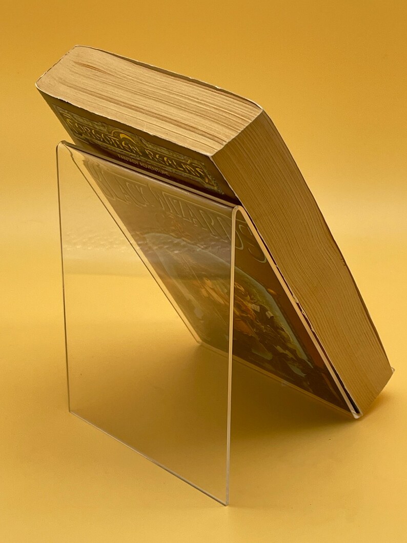 Op de afbeelding: Een heldere acryl boekenstandaard met een hardcover boek. Het boek staat schuin, waardoor de kaft en pagina's zichtbaar zijn. De standaard is transparant, waardoor de kaft van het boek zichtbaar is. De achtergrond is effen geel.