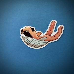 May include: A sticker featuring a cartoon illustration of a creature with an open mouth, resting on a striped surface, and a hand. The creature is orange and black, and the hand is flesh-toned. The sticker has a white border.