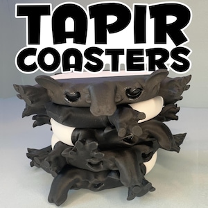 May include: A stack of black tapir-shaped coasters with white and black accents. The coasters have a unique design, featuring tapir faces and feet. The text "TAPIR COASTERS" is displayed in bold, black letters with a white outline.