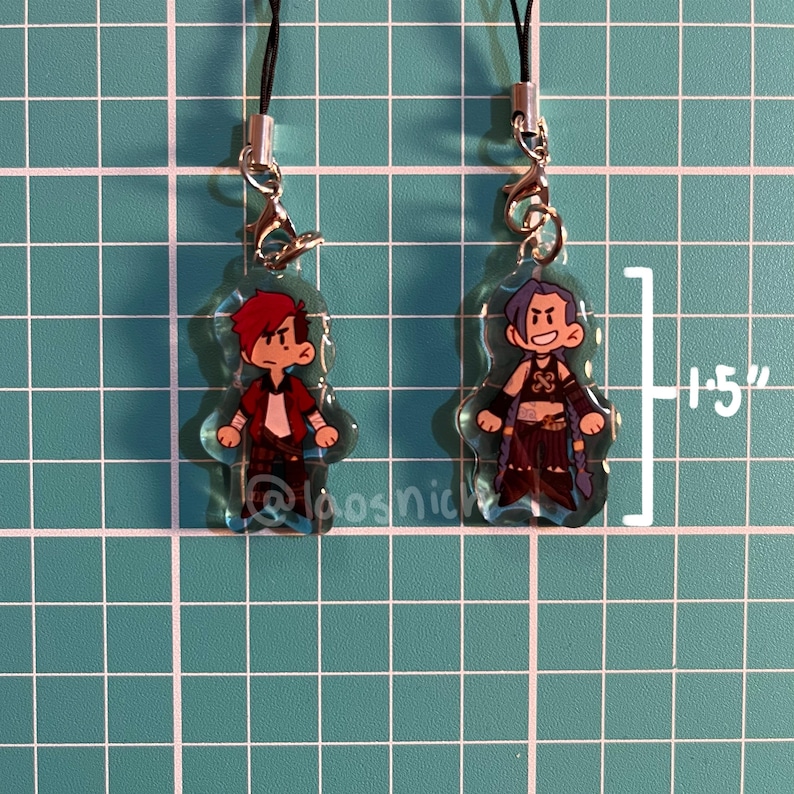 K&ouml;nnte beinhalten: Zwei transparente Acryl-Anh&auml;nger mit Cartoon-Charakter-Designs. Einer zeigt einen Charakter mit roten Haaren und einem wei&szlig;en Hemd unter einer roten Jacke. Der andere hat blaue Haare und ein dunkles Outfit. Jeder Anh&auml;nger ist etwa 3,8 cm hoch.
