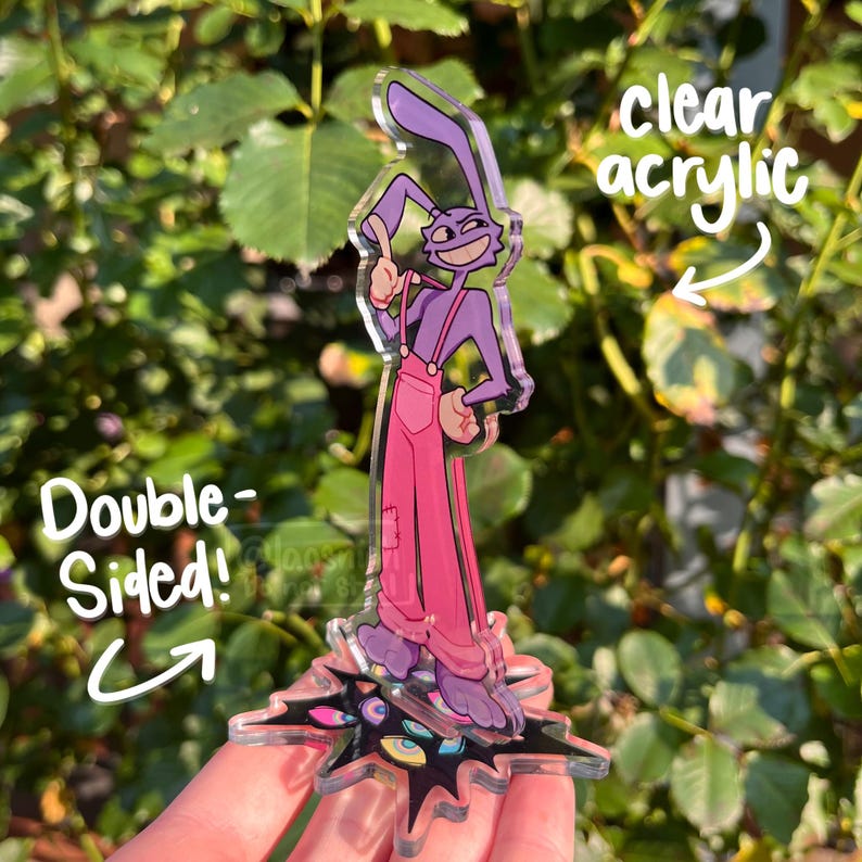 May include: A double-sided clear acrylic charm featuring a purple rabbit character wearing pink overalls. The charm has a starburst base with colorful eye designs. Text on the charm reads "clear acrylic" and "Double-Sided!"