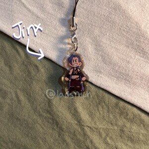 K&ouml;nnte beinhalten: Ein klarer Acryl-Anh&auml;nger mit einer Cartoon-Figur mit blauen Haaren und einem schelmischen Gesichtsausdruck. Der Anh&auml;nger ist an einer schwarzen Kordel und einem Metallverschluss befestigt. Das Wort "Jinx" ist in einer verspielten Schrift geschrieben.