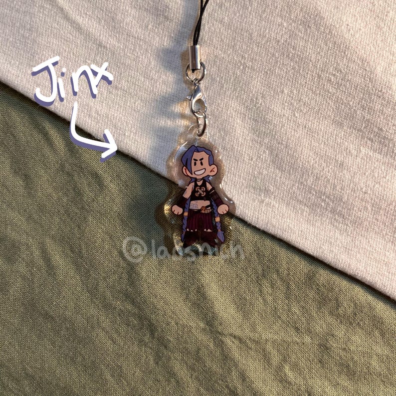 K&ouml;nnte beinhalten: Ein klarer Acryl-Anh&auml;nger mit einer Cartoon-Figur mit blauen Haaren und einem schelmischen Gesichtsausdruck. Der Anh&auml;nger ist an einer schwarzen Kordel und einem Metallverschluss befestigt. Das Wort "Jinx" ist in einer verspielten Schrift geschrieben.