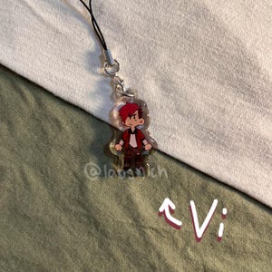 K&ouml;nnte beinhalten: Ein klarer Acryl-Anh&auml;nger mit einer Cartoon-Figur mit roten Haaren, einer roten Jacke und braunen Hosen. Der Anh&auml;nger ist an einer schwarzen Kordel mit einem silbernen Verschluss befestigt. Das Wort "Vi" ist wei&szlig; mit einem Pfeil geschrieben.