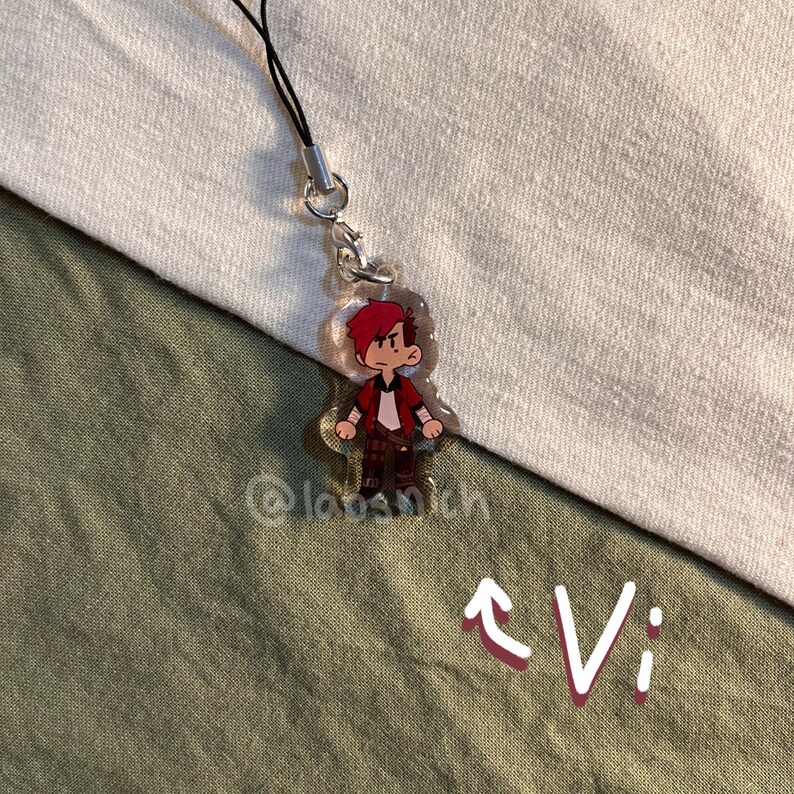 K&ouml;nnte beinhalten: Ein klarer Acryl-Anh&auml;nger mit einer Cartoon-Figur mit roten Haaren, einer roten Jacke und braunen Hosen. Der Anh&auml;nger ist an einer schwarzen Kordel mit einem silbernen Verschluss befestigt. Das Wort "Vi" ist wei&szlig; mit einem Pfeil geschrieben.