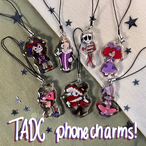 Puede incluir: Colección de siete colgantes de teléfono de acrílico transparente con personajes de dibujos animados. Cada colgante tiene un diseño único con colores vibrantes, incluyendo un bufón, un rey, un esqueleto y una niña con un vestido morado. El texto "TADC phone charms!" está en la parte inferior.
