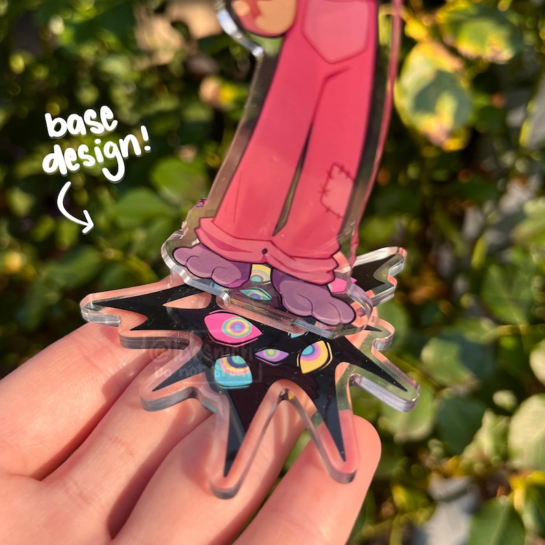 May include: A clear acrylic charm featuring a cartoon character in pink overalls and purple feet, standing on a black star-shaped base with colorful eye designs. The text "base design!" is written in white.