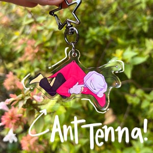 Könnte beinhalten: Ein klarer Acryl-Schlüsselanhänger mit einer Cartoonfigur in einem roten Mantel und einer schwarzen Hose, mit dem Text "Ant Tenna!". Der Schlüsselanhänger hat eine silberne sternförmige Oberseite und einen silbernen Verschluss.