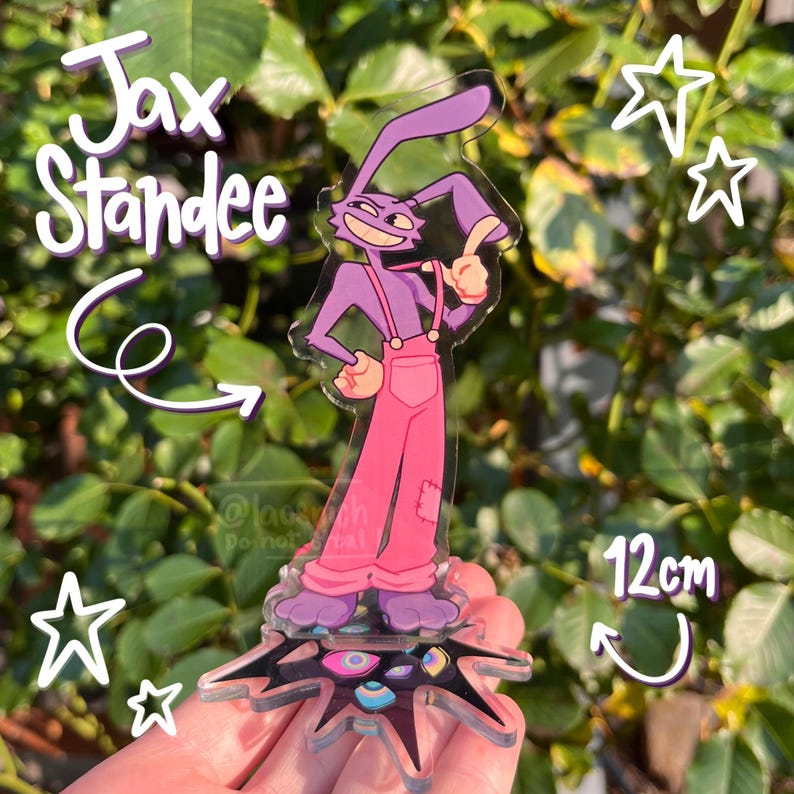 May include: A Jax Standee acrylic figure featuring a purple rabbit character wearing pink overalls. The figure is approximately 12cm tall and has a unique base. The text "Jax Standee" is visible. The background is a green leafy plant.