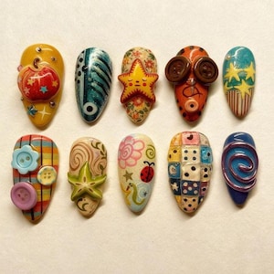 Könnte beinhalten: Zehn künstliche Fingernägel mit individuellen Designs. Jeder Nagel zeigt ein anderes Muster, darunter ein Kürbis, ein Seestern, Knöpfe und Würfel. Die Farben umfassen Gelb, Blau, Orange und Rosa, mit verschiedenen Mustern und Verzierungen.