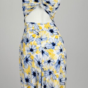 Könnte beinhalten: Ein ärmelloses Kleid mit weißem Grund und einem floralen Muster in Blau- und Gelbtönen. Das Oberteil hat ein gedrehtes Knotendetail in der Mitte des Rückens, das einen Ausschnitt über der taillierten Passform und dem fließenden Rock erzeugt.
