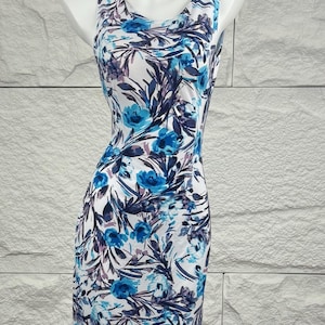 May include: Sleeveless sheath dress with a floral pattern. The dress has a white background with blue and purple floral designs. The dress is displayed on a mannequin against a textured white wall.