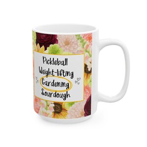 Puede incluir: Taza de cerámica blanca con un diseño floral y las palabras "Pickleball, Weight-lifting, Gardening, Sourdough" en texto negro. La palabra "Gardening" está resaltada en un círculo amarillo. Pequeños gráficos de corazón a izquierda y derecha.