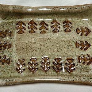 Puede incluir: Fuente rectangular de cerámica con un esmalte moteado en tonos verdes y marrones terrosos. La fuente presenta un patrón repetitivo de diseños estilizados de hojas y flechas en un color marrón más oscuro. Los bordes están ligeramente elevados.