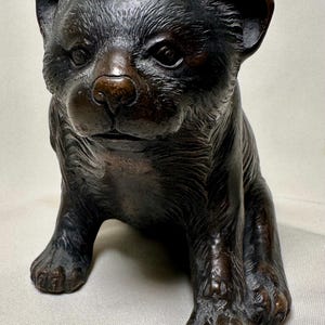 May include: A bronze sculpture of a seated bear cub. The cub has detailed fur texture and is depicted with a serious expression. The sculpture is a dark brown color and has a polished finish. The bear cub is a decorative item.