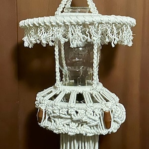 May include: A handmade macrame lamp with a clear glass globe. The white macrame features intricate knotting and fringe details. The lamp has a wooden accent and a hanging cord. The overall design is bohemian and decorative.