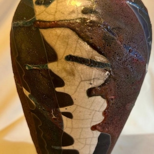 May include: A handcrafted ceramic vase with a unique, abstract design. The vase features a combination of dark brown, cream, and black colors with accents of green and blue. The surface has a textured, crackled appearance, adding to its artistic appeal.