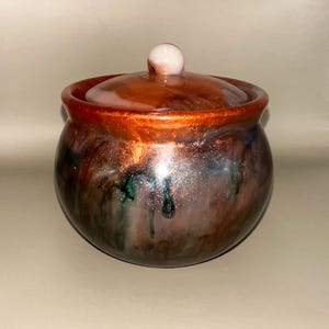 Könnte beinhalten: Ein kleiner, runder Topf mit Deckel und glänzendem Finish. Der Topf ist hauptsächlich braun mit grünen und silbernen Streifen und einem kupferfarbenen Rand. Der Deckel hat einen kleinen, runden, weißen Knopf. Der Topf scheint handgefertigt zu sein.