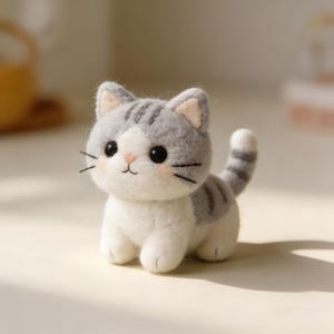 Puede incluir: Una figura de gato de fieltro hecha a mano en gris y blanco. El gato tiene ojos negros, bigotes y una nariz rosa. Las orejas y la cola están a rayas. Este adorable gato es un objeto decorativo encantador.