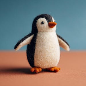 May include: A handcrafted felt penguin figurine with a white body, black head and wings, and orange beak and feet. The penguin has a charming, friendly expression and is set against a blue and peach background.
