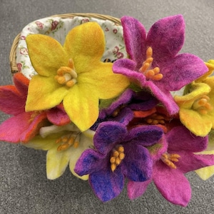 May include: A collection of colorful felt flowers in a woven basket. The flowers are in shades of yellow, pink, purple, and orange. The basket has a floral patterned lining. The flowers have detailed stamens and petals.