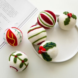 Peut inclure: Ornements de Noël faits à la main. Les boules sont blanches, rouges et vertes, avec des motifs variés tels que des rayures, des feuilles de houx et des baies. Chaque ornement est muni d'une ficelle pour être suspendu.